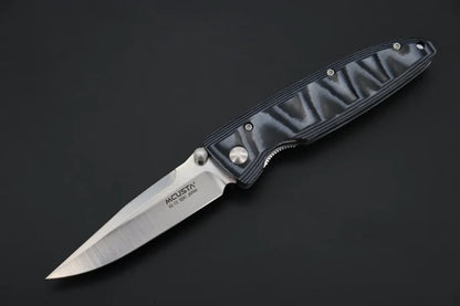 A folding pocket knife with a satin-finished silver blade and a textured blue/grey handle, shown open against a dark gray background. The blade is engraved with the brand name 'MCUSTA'