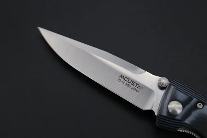 A close-up of an MCUSTA folding pocket knife with a satin-finished silver blade and a textured black handle, set against a dark black background.