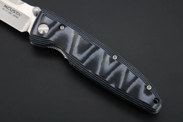 A close-up of an MCUSTAf olding pocket knife with a textured black handle and a silver blade, set against a dark background.