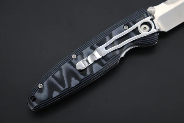 A close-up of a folding knife with a textured black handle scale and a silver pocket clip, shown against a dark background.