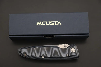 An MCUSTA folding pocket knife with a textured black handle and silver metal hardware, displayed below its dark navy blue presentation box on a dark background.