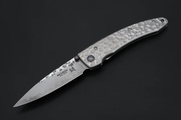 A folding pocket knife with a hammered silver handle and a patterned steel blade, shown open against a dark background. The blade features the brand name 'MCUSTA' and a logo.
