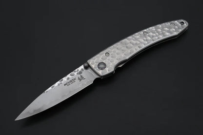 A folding pocket knife with a hammered silver handle and a patterned steel blade, shown open against a dark background. The blade features the brand name 'MCUSTA' and a logo.