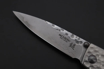 A close-up of a MCUSTA folding-blade knife with a hammered Damascus steel blade and textured handle, set against a dark background.