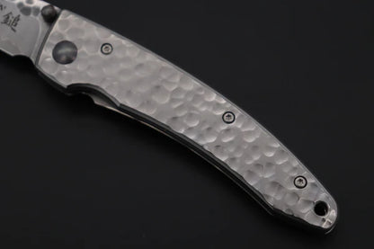 A close-up of a folding pocket knife with a hammered metal handle and a partially visible blade, photographed on a dark background.