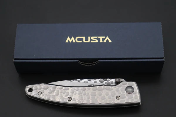 A silver folding pocket knife with a hammered texture and a silver blade, displayed in front of its dark navy blue presentation box with gold 'MCUSTA' branding.
