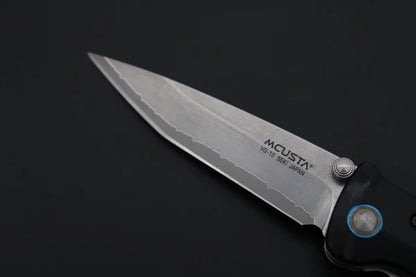 A close-up of a MCUSTA folding pocket knife with a silver steel blade and a black handle, set against a dark black background.