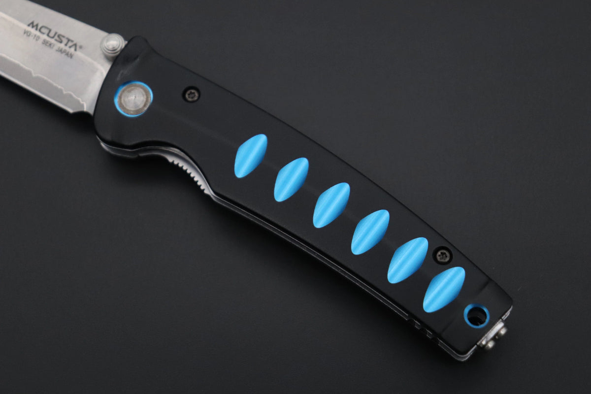 A close-up of an MCUSTA folding knife with a black handle featuring bright blue oval inlays and a silver stainless steel blade, set against a dark gray background.