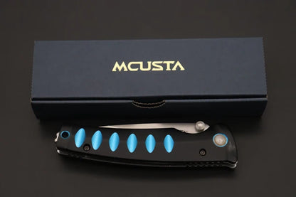 A black folding pocket knife with bright blue accents and a silver blade, displayed in front of its dark navy blue Mcusta brand box on a dark background.