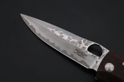 A close-up of a folding-blade knife with a hammered Damascus steel blade and a dark brown wooden handle, shown on a dark background. The blade is branded with 'MCUSTA'.