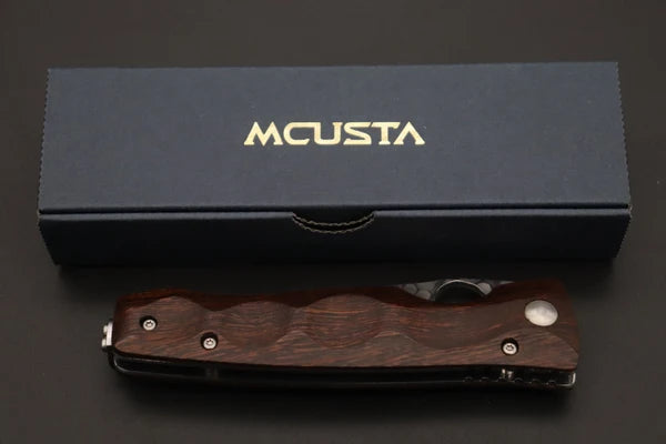 A closed folding pocket knife with a dark wood handle and silver hardware, displayed in front of its dark navy blue presentation box with gold 'MCUSTA' branding.