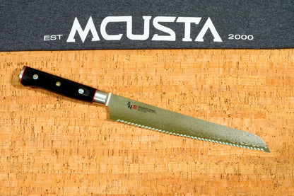A long Japanese-style bread knife with a black handle and serrated blade, placed diagonally on a cork cutting board. A dark gray banner at the top features the white text 'EST MCUSTA 2000'.