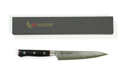 A Mcusta Petty knife with a textured steel blade and a black handle, displayed below its dark gray presentation box on a white background.
