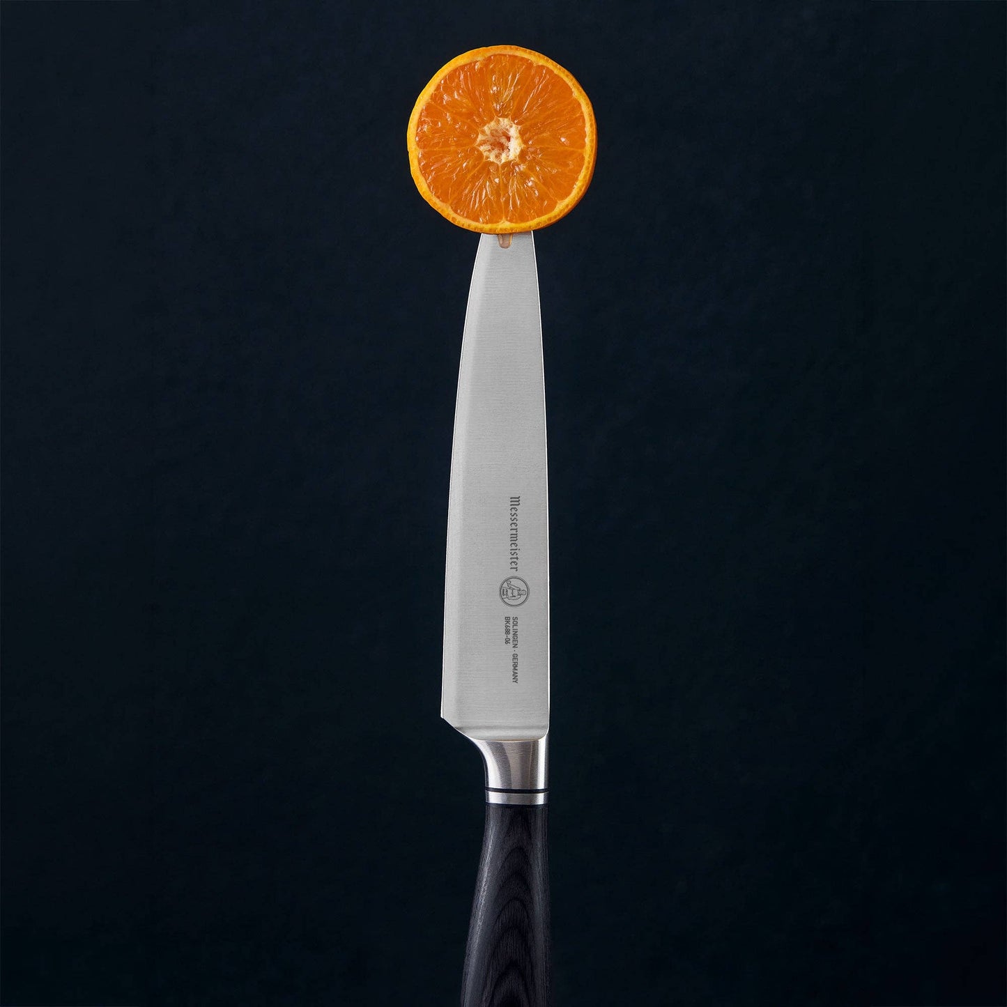 A Blacksmith utility knife with a silver blade and black handle, balanced on its tip with a sliced orange resting on the blade, set against a dark textured background.