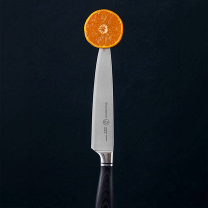 A Blacksmith utility knife with a silver blade and black handle, balanced on its tip with a sliced orange resting on the blade, set against a dark textured background.
