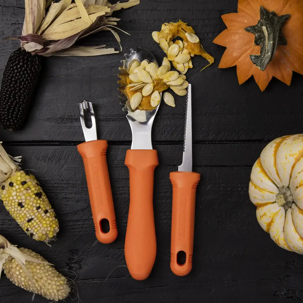 Three orange-handled tools on a dark surface with pumpkins and corn ears.