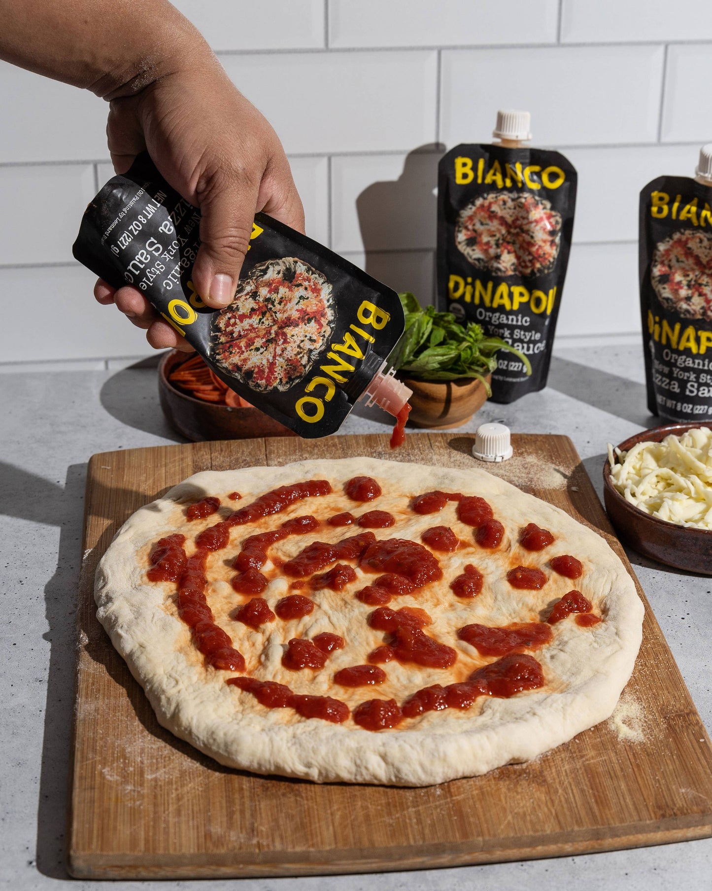 Person drizzling pizza sauce on a pizza with Bianco products in the background.