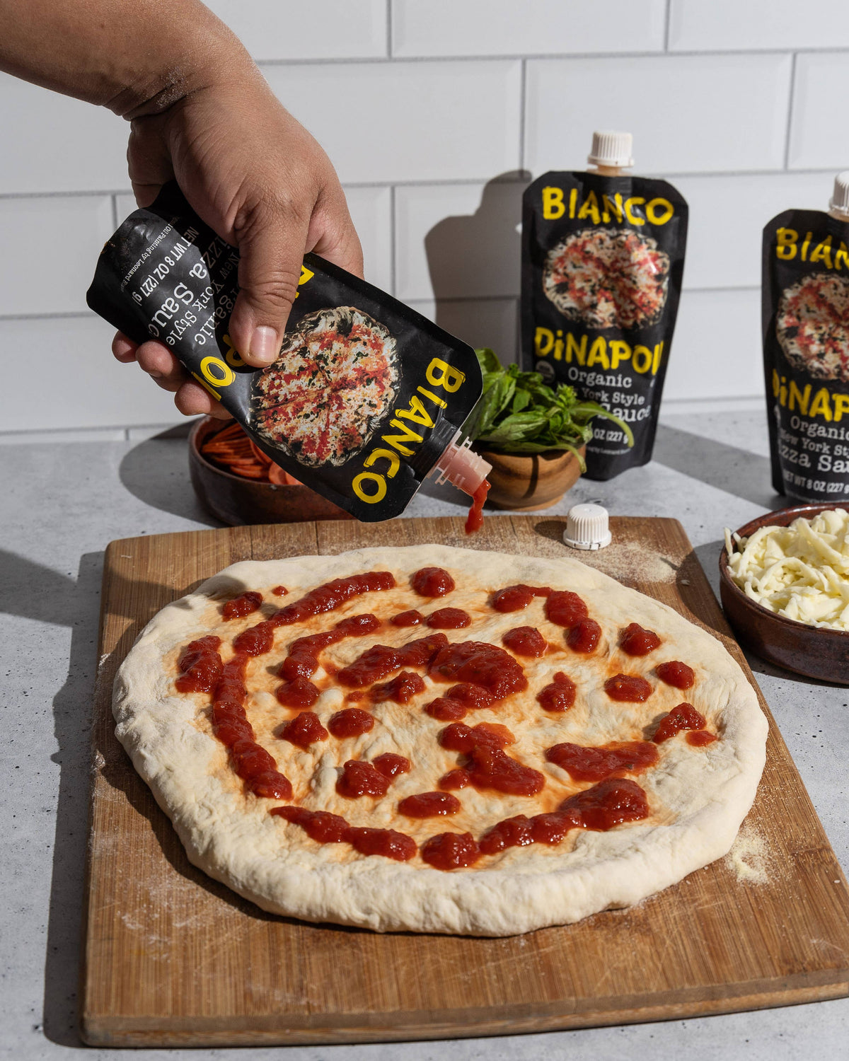 Person drizzling pizza sauce on a pizza with Bianco products in the background.
