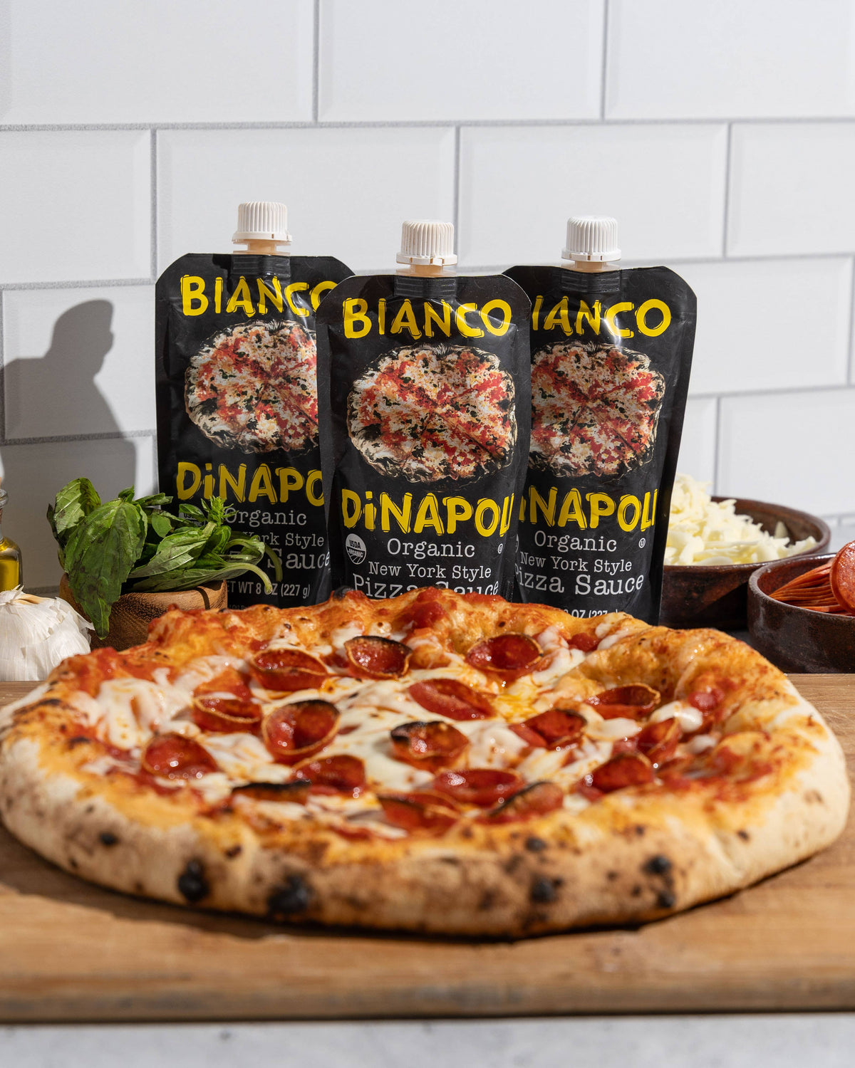 Pizza with pepperoni on a wooden board with Bianco Dinapoli pizza sauce packages in the background.