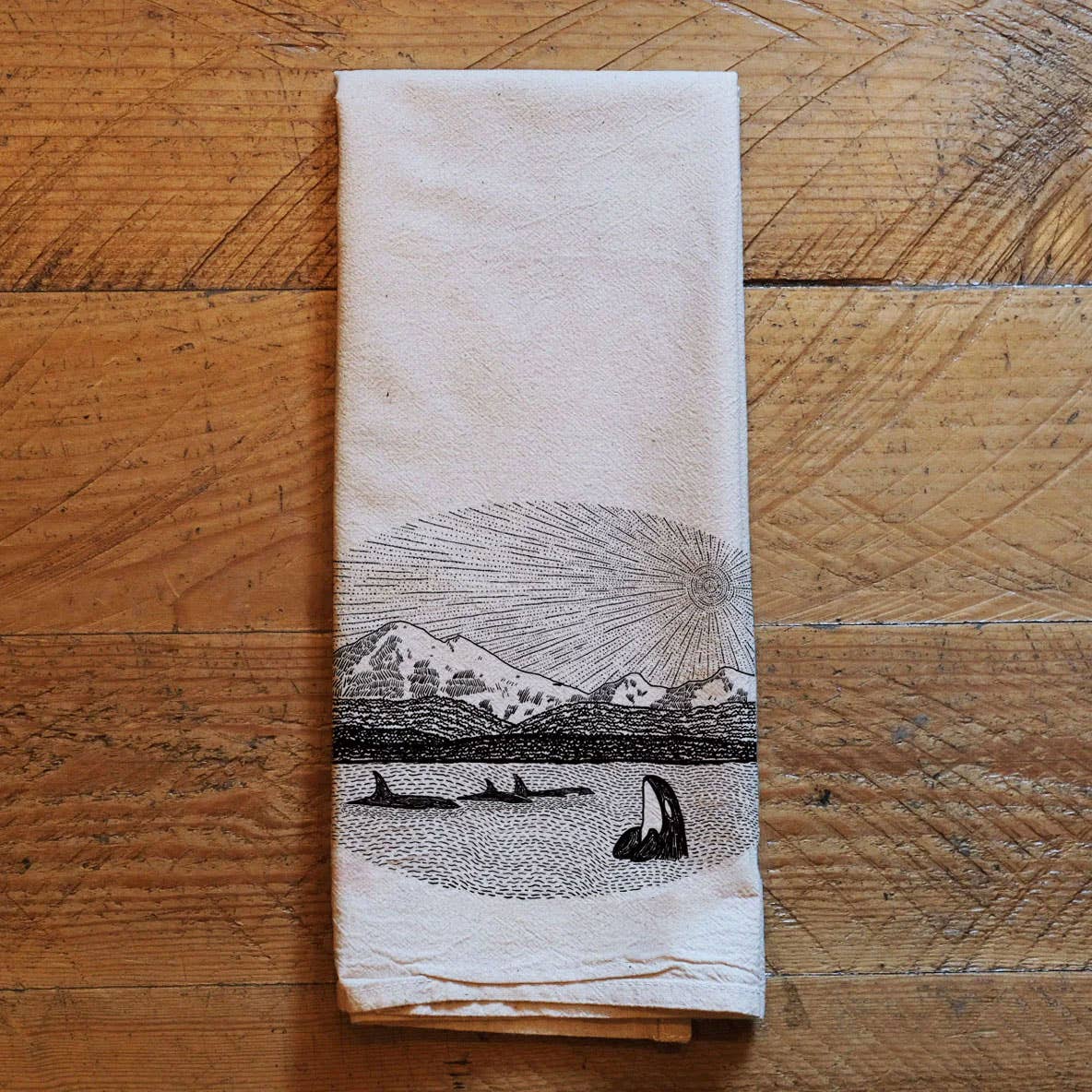 Folded towel with a black and white illustration of mountains and whales on a wooden surface