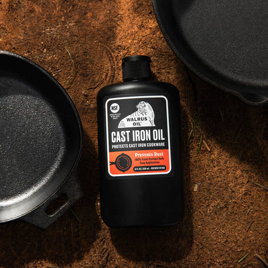 Bottle of Walrus Oil Cast Iron Oil with a cast iron skillet on a rustic background