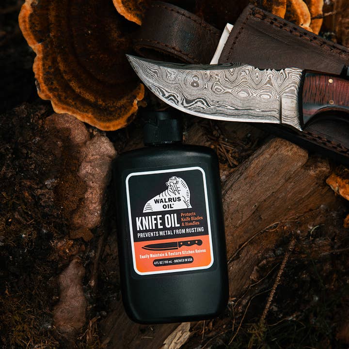 A bottle of Walnut Oil Knife Oil placed on rustic wood logs, with a partially unsheathed fixed-blade knife and a brown leather sheath behind it. The label reads Walrus Oil Knife Oil.