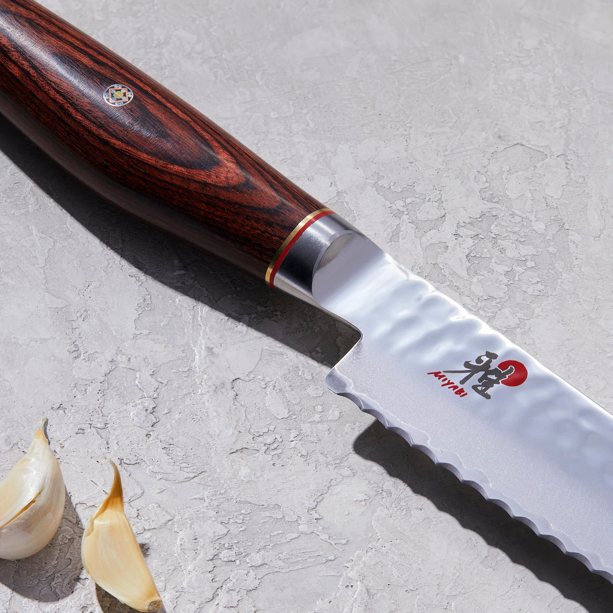Closeup of Miyabi Artisan Bread Knife with Miyabi logo and rosewood handle with mosaic pin on textured white background with cloves of garlic