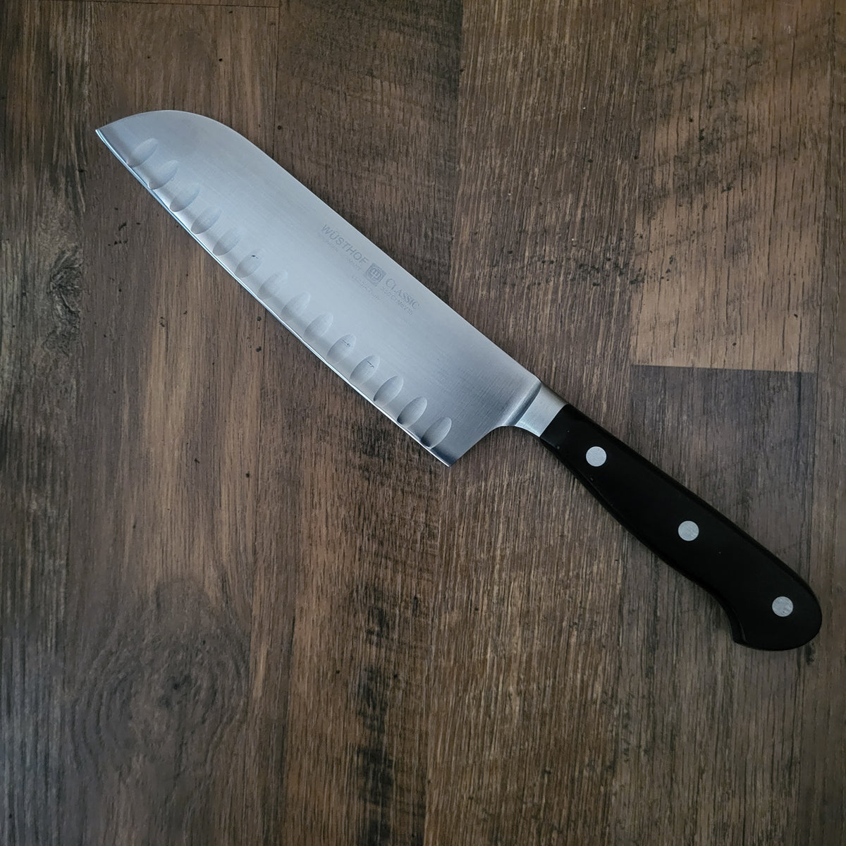 A Santoku-style kitchen knife with a silver steel blade and a black handle, placed diagonally on a dark brown wood grain surface.